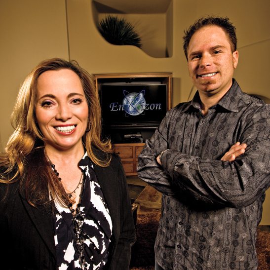 Heidi and Mike standing in front of a TV with their logo on the screen