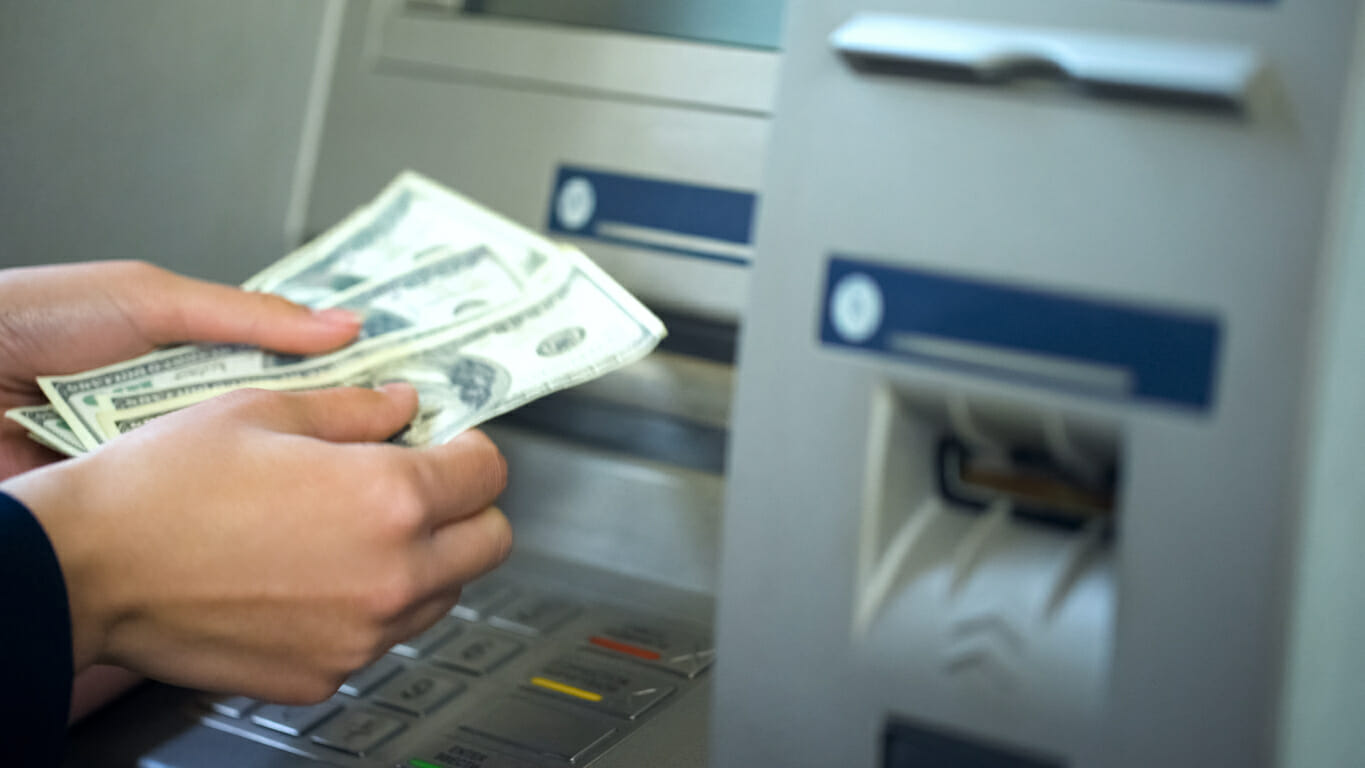 Person's hand holding a stack of U.S. dollars in front of an ATM.