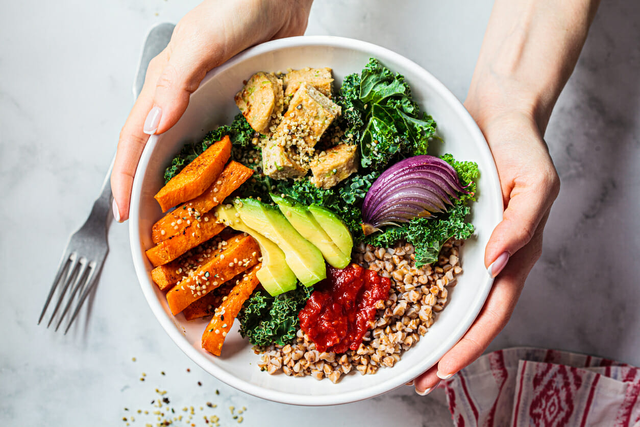 Two hands hold a bowl of avocado, quinoa, sweet potatoes and assorted veggies.