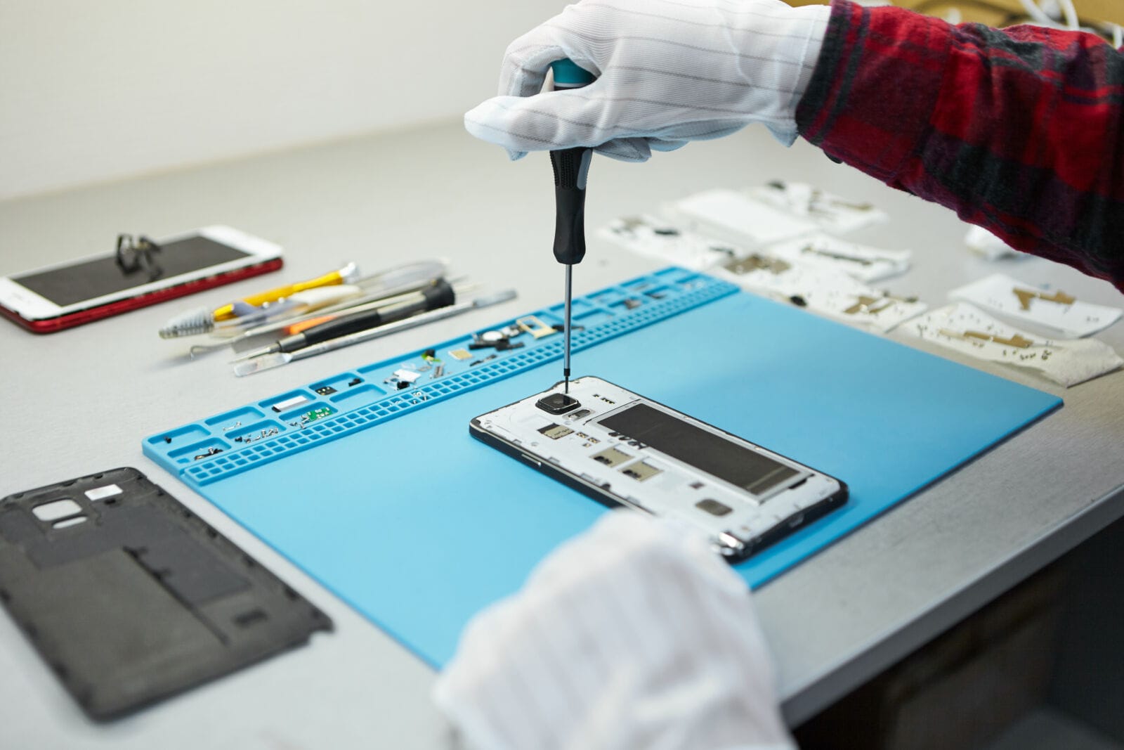 Cropped shot of male technician in antistatic gloves using screwdriver to disassemble borken mobile phone, going to repair motherboard, sitting at his workplace in laboratory with needed equipment.
