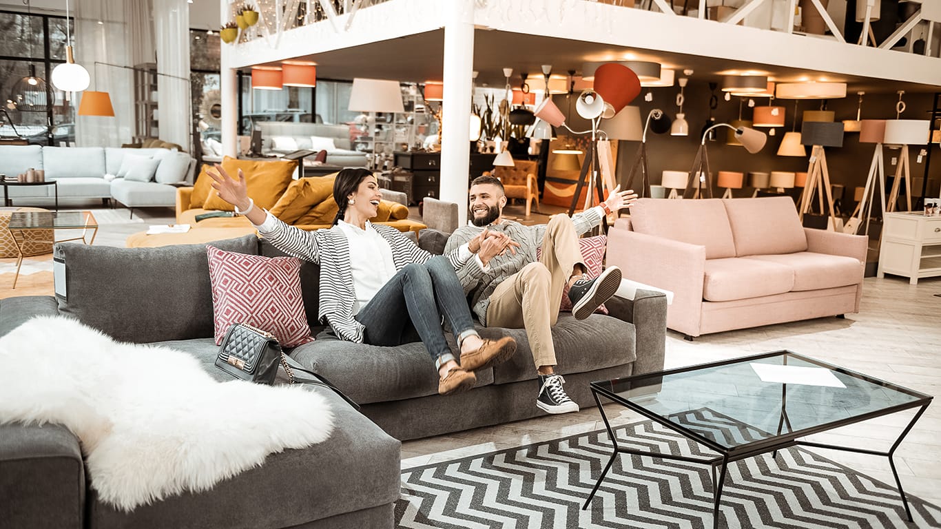 After long day. Cheerful laughing couple behaving childishly in furniture showroom while fooling around on grey couch.