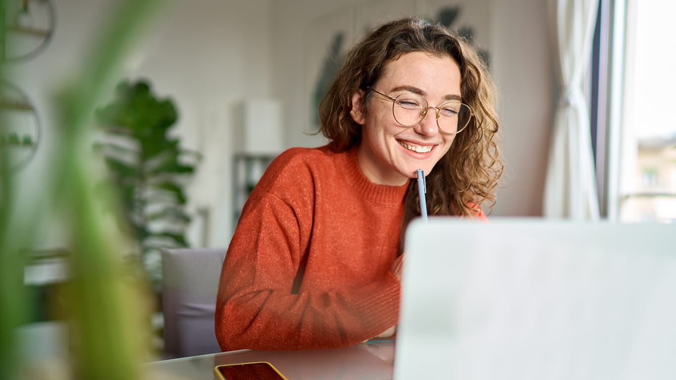 Happy young woman using laptop sitting at desk writing notes while watching webinar, studying online, looking at pc screen learning web classes or having virtual call meeting remote working from home.