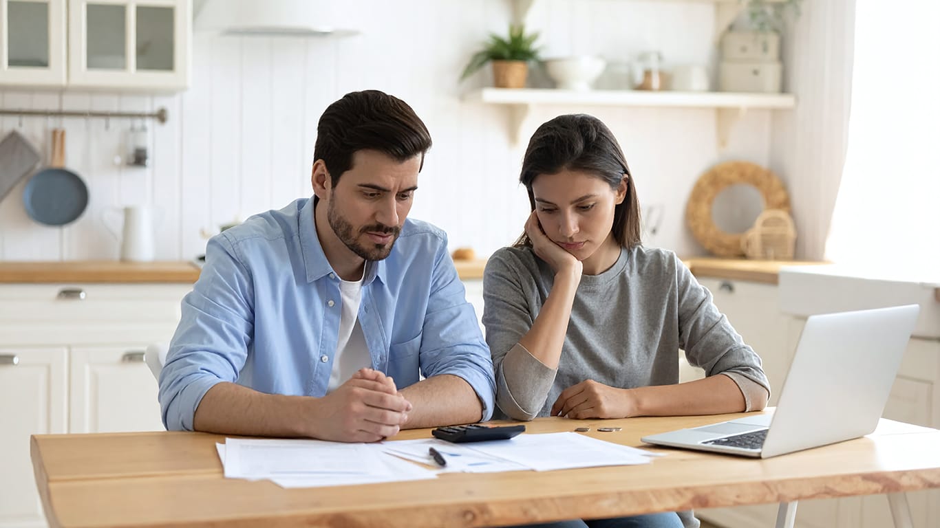 A concerned couple sits in their bright kitchen, meticulously reviewing bills and financial documents. This image captures the stress of budgeting, debt management, and household finance.