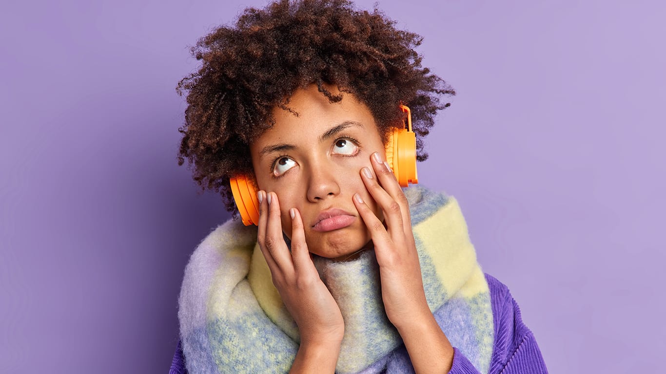 Headshot of curly haired woman feels bored as listens monotonous song has displeased dull uninterested expression wears stereo headphones on ears wears fashionable winter clothes.