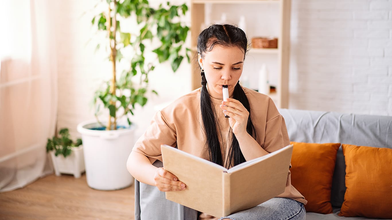 A beautiful young woman uses a disposable electronic cigarette on the couch at home reading a book. vaping concept.