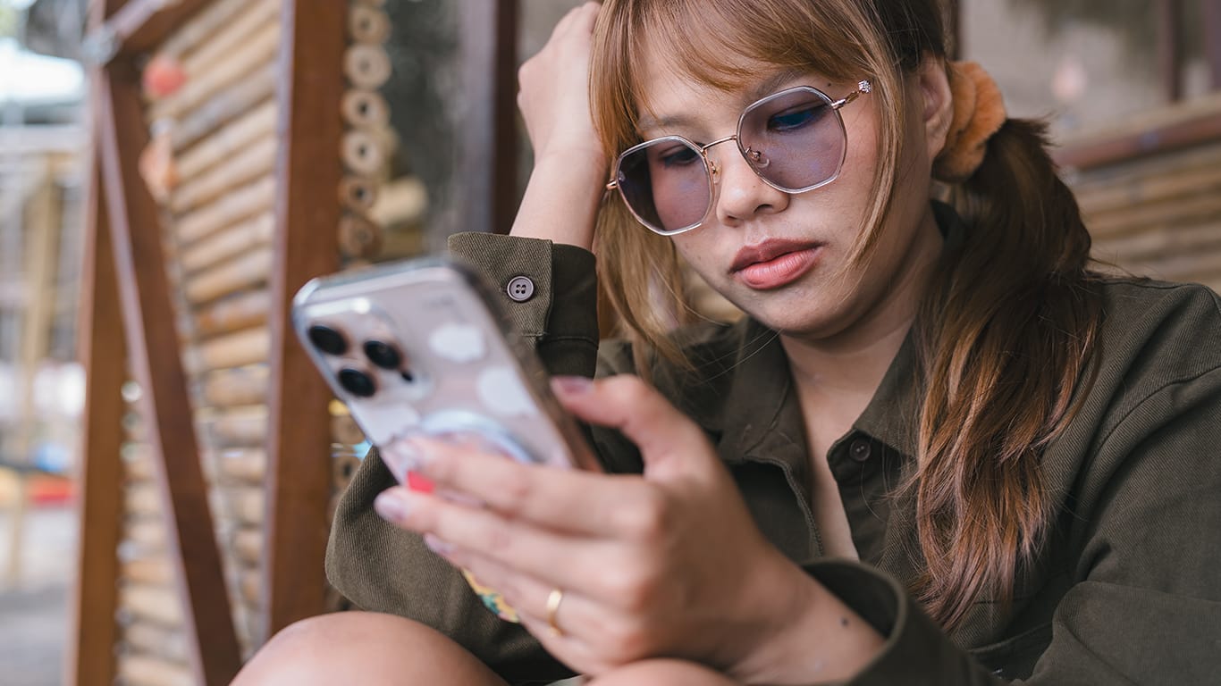 Negative emotion and digital stress concept worried Asian woman with sunglasses looking at smartphone reading bad news or dealing with cyberbullying problem during outdoor summer vacation.