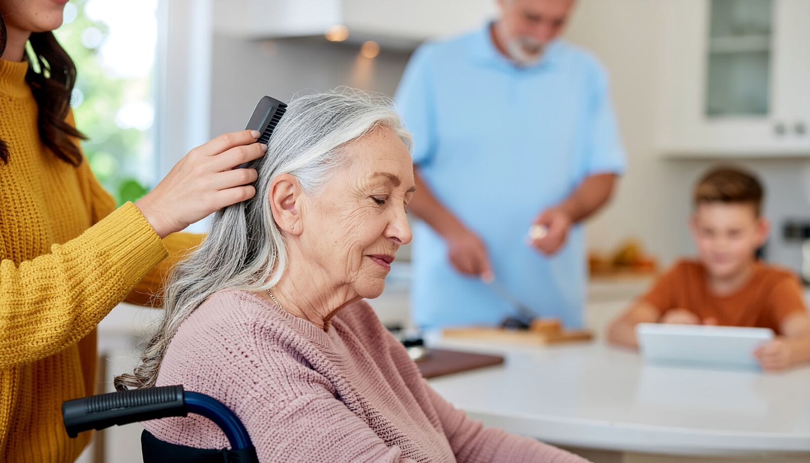 A family member combs the hair of an elderly woman in a wheelchair in a kitchen.