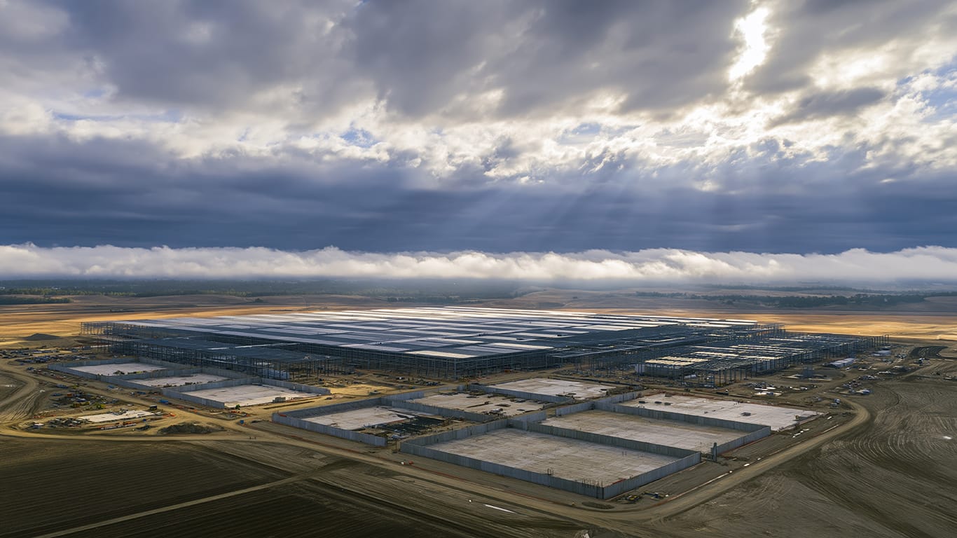 Aerial perspective of a massive data center with solar panels, situated in an expansive landscape under a cloudy sky with sunlight breaking through.