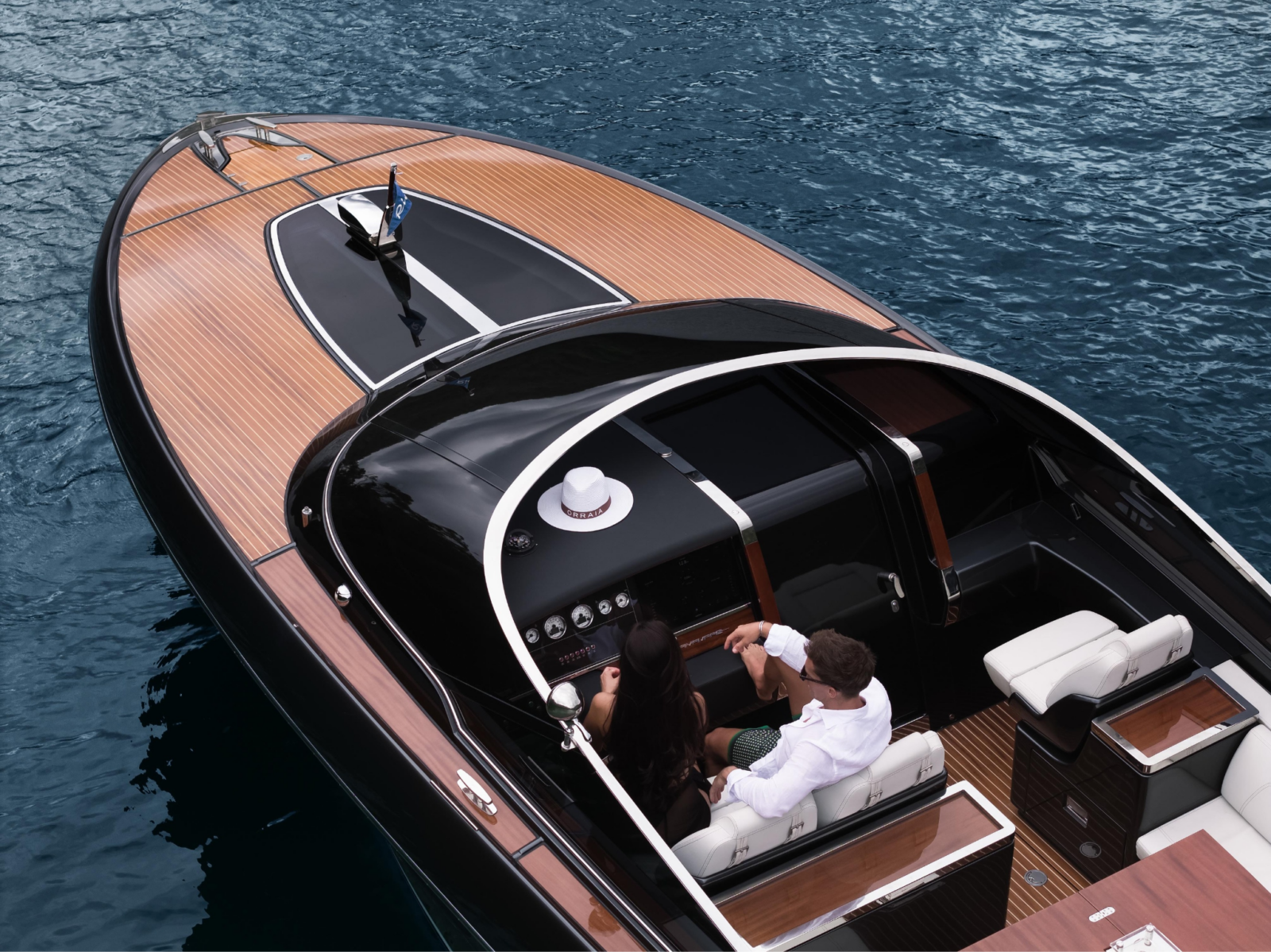 A couple seated aboard a sleek luxury yacht, viewed from above as the vessel moves across deep blue water.