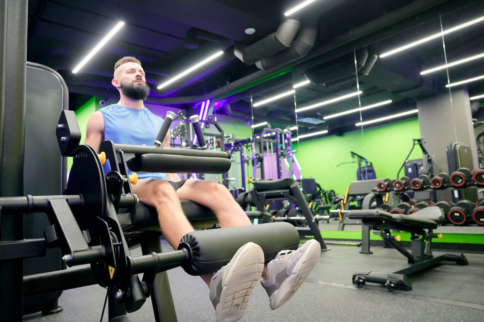 Athletic man performs leg extensions on a gym machine inside a modern fitness center at night, focusing on his quad muscles during a regular workout routine.