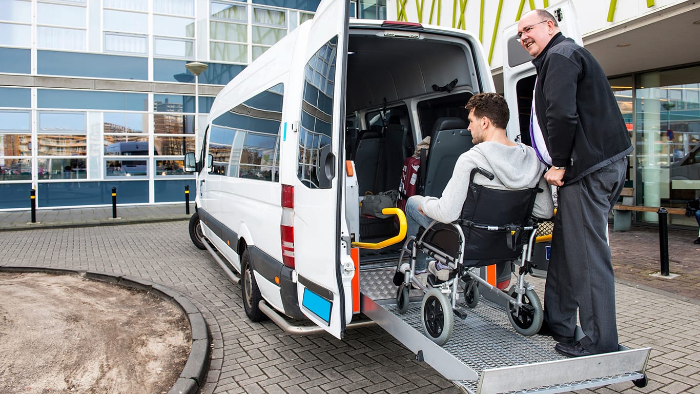 The driver of a wheel chair taxi, helping a disabled man in a wheel chair, using the lift in the back of his mini van.