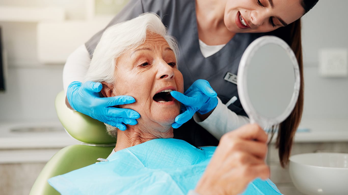 Dentist, senior woman and consulting with mirror for teeth cleaning, dental implant or oral hygiene. Inspection, healthcare or doctor with old patient in clinic for mouth exam or root canal treatment.