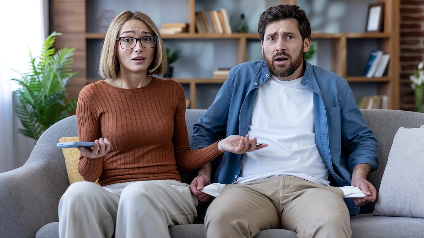 Frustrated young couple sitting on a sofa at home, facing financial difficulties and showing empty pockets, feeling stressed about bankruptcy and money problems.